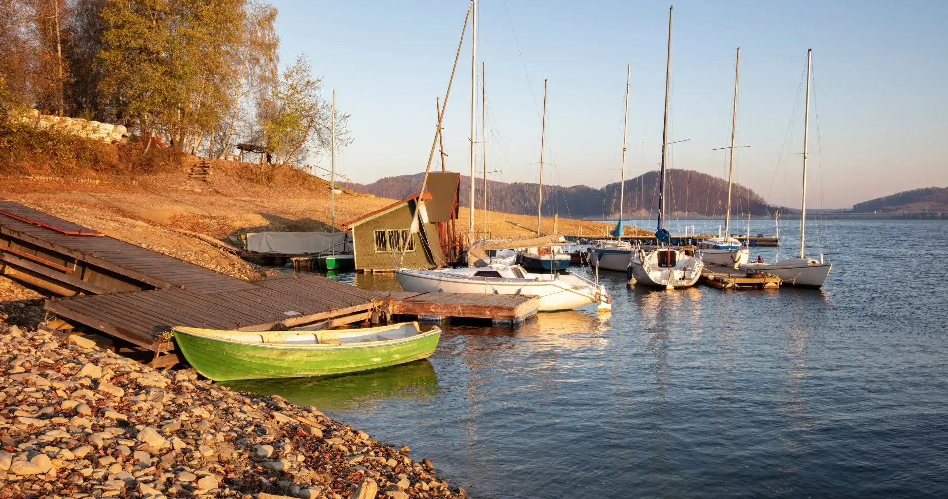 Przystań żeglarska nad brzegiem jeziora w Bieszczadach, otoczona jesiennym krajobrazem. Łodzie i jachty zacumowane przy drewnianych pomostach, w tle złote liście i pagórkowaty teren.