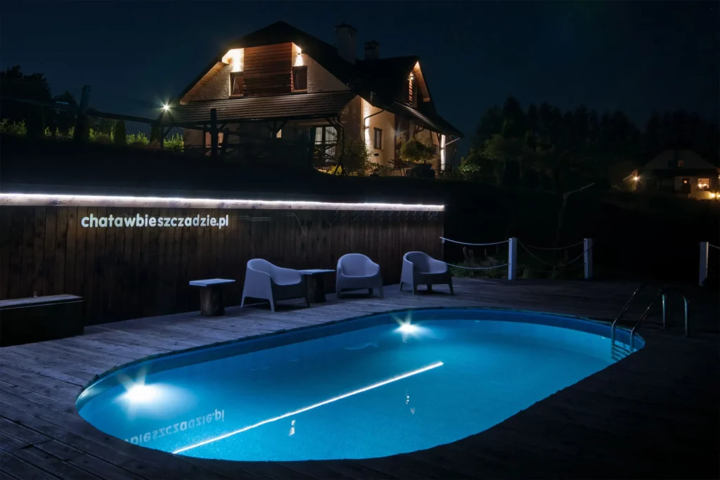 Nocny widok na basen z podświetloną wodą na drewnianym tarasie, z chatą w tle oświetloną ciepłymi światłami.