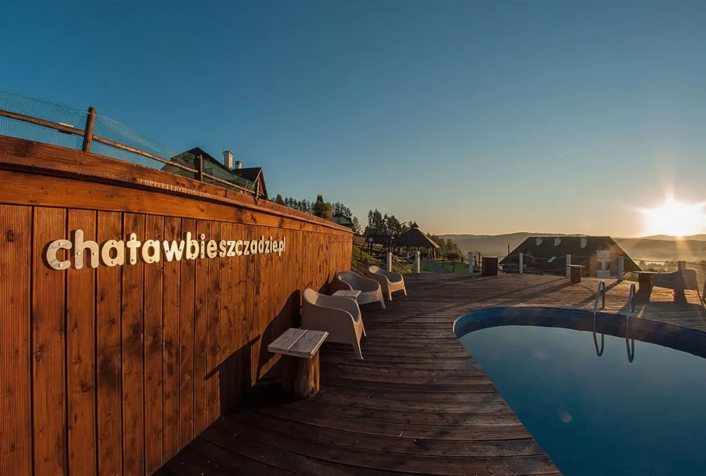 Drewniany taras z basenem i leżakami w Bieszczadach, na tle widoczny dom i wschód słońca.