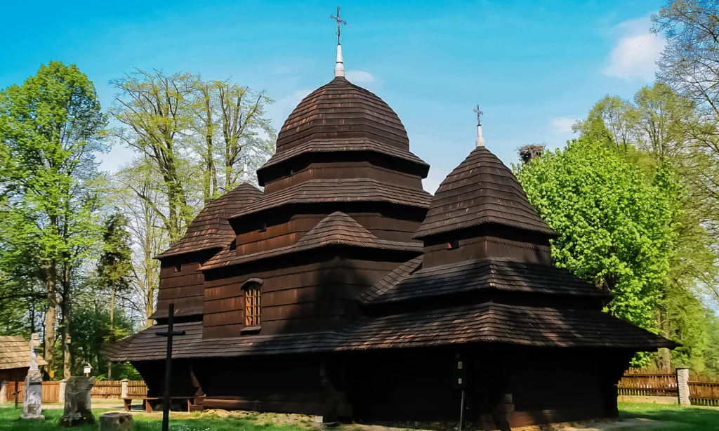 Zabytkowa drewniana cerkiew w Sanoku, o charakterystycznej architekturze, otoczona zielonymi drzewami, na tle błękitnego nieba w słoneczny dzień.