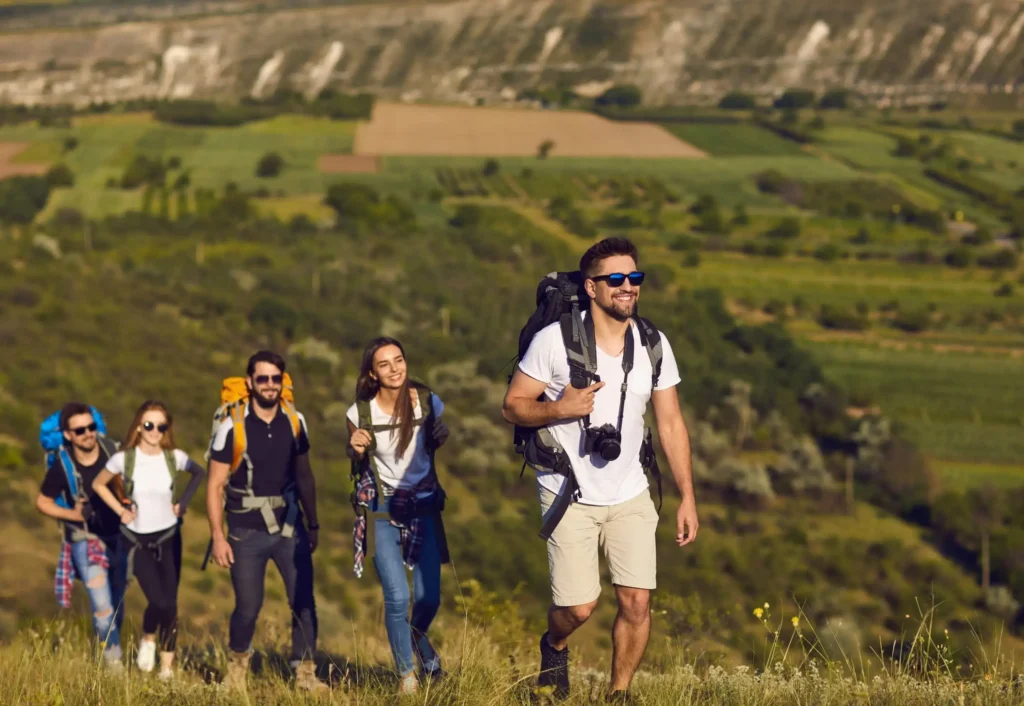 Grupa młodych ludzi wędrująca razem po zielonych wzgórzach, uśmiechnięta i z plecakami.