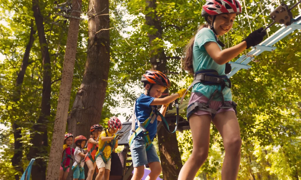 Dzieci w kaskach i uprzężach bawią się w parku linowym w Bieszczadach. Uśmiechnięte pokonują przeszkody na trasie wśród drzew.