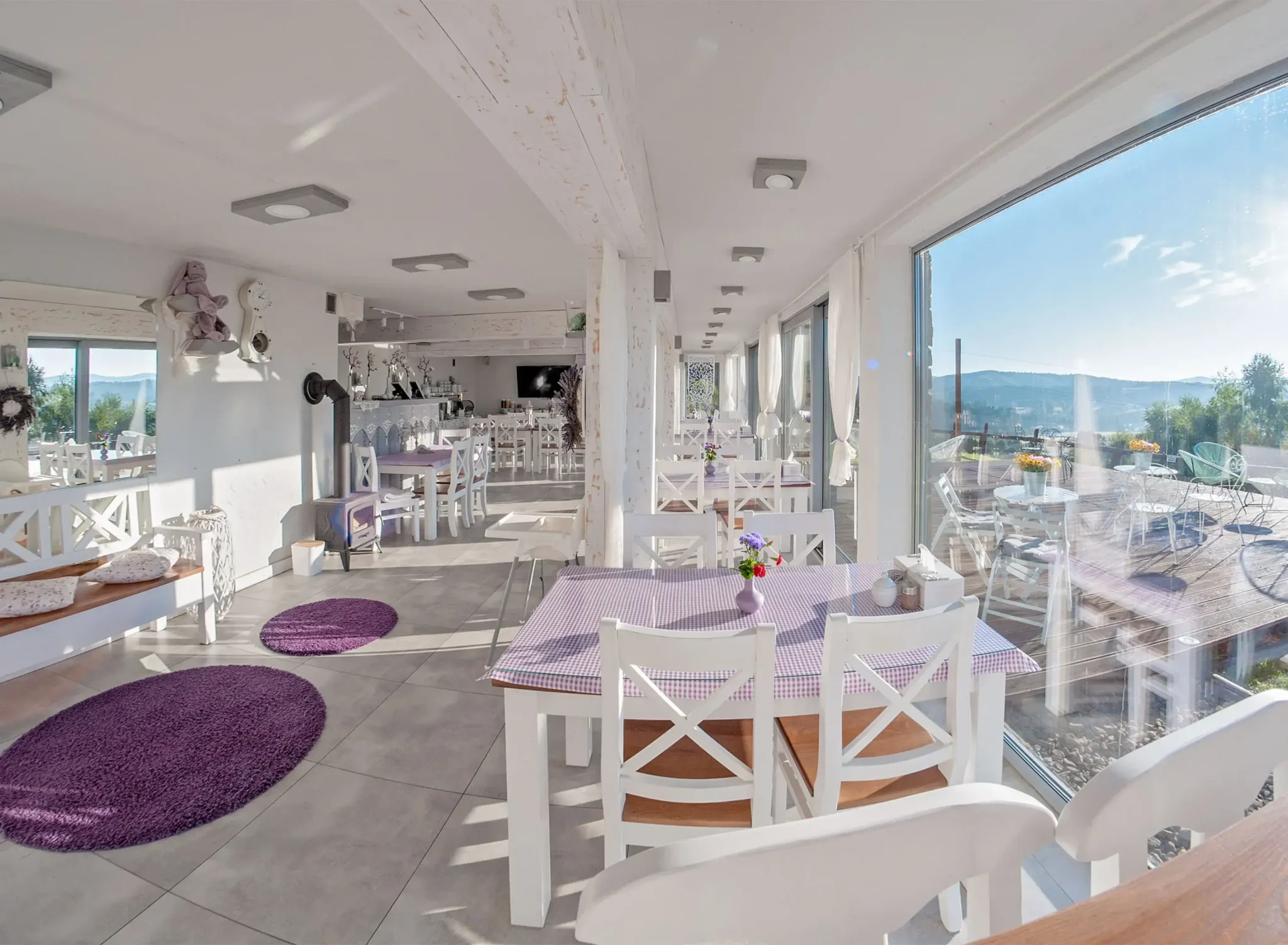 Jasna sala restauracyjna w Chacie w Bieszczadzie, z białymi meblami, fioletowymi dodatkami i dużymi oknami wychodzącymi na taras z widokiem na góry.