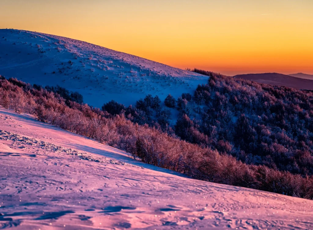 Widok na ośnieżone zbocze Wielkiej Rawki w Bieszczadach podczas zachodu słońca, z pomarańczowym niebem kontrastującym z różowymi i niebieskimi odcieniami śniegu. Idealne miejsce na zimowe wędrówki i podziwianie górskiej panoramy.
