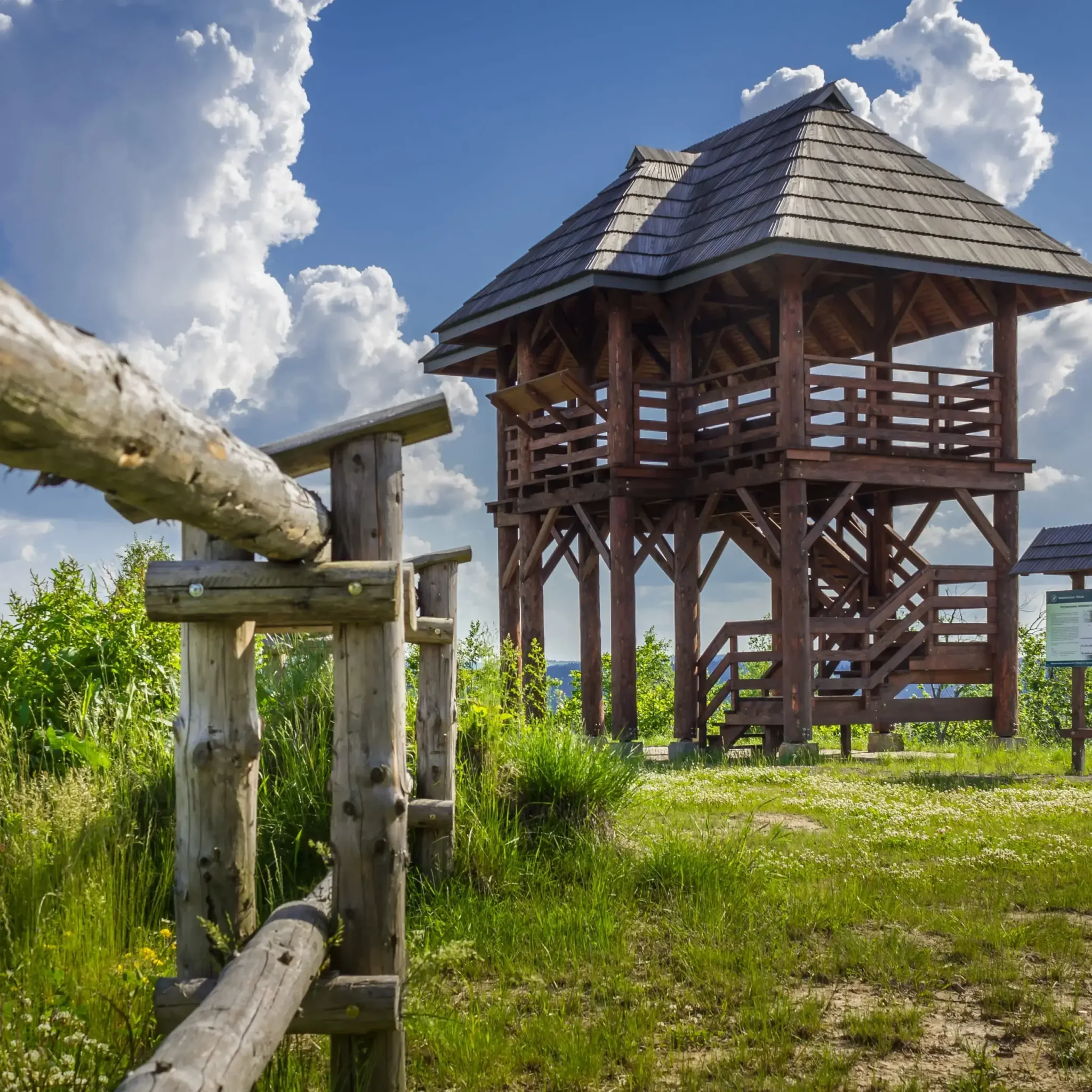 Drewniana wieża widokowa otoczona zielenią na tle błękitnego nieba z malowniczymi chmurami. Drewniane ogrodzenie w pierwszym planie podkreśla rustykalny charakter krajobrazu.
