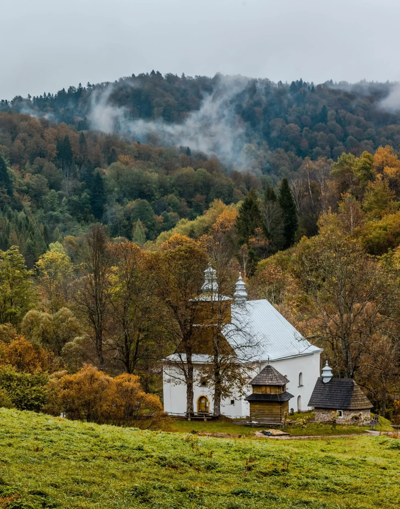 Zabytkowa cerkiew w Łopience, otoczona drzewami w jesiennych kolorach i mglistym krajobrazem Bieszczad. Malownicza sceneria historycznego miejsca.