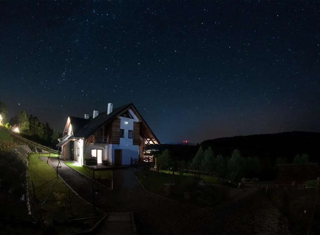 Widok na drewnianą chatę w Bieszczadach nocą, oświetloną światłem domowym, z gwiaździstym niebem w tle.