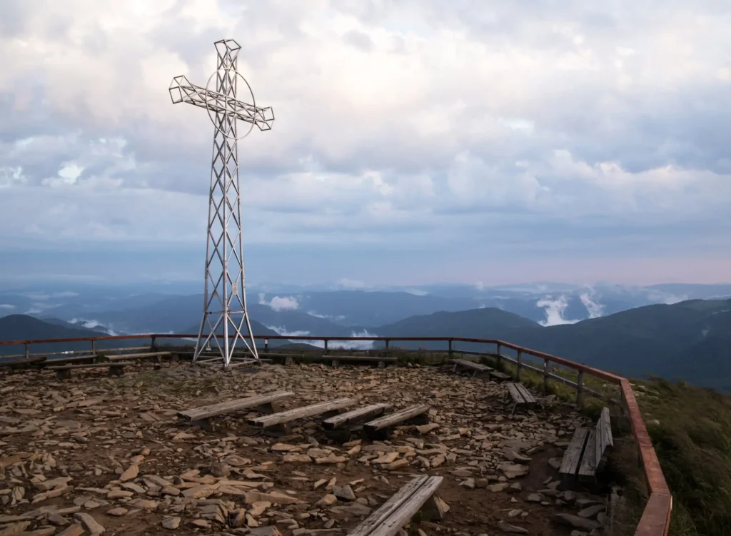 Metalowy krzyż na szczycie Tarnicy, najwyższej góry Bieszczad, otoczony kamienistym terenem i ławkami dla turystów. W tle rozciągają się majestatyczne widoki górskich szczytów i poranne mgły. Idealne miejsce dla miłośników trekkingu i natury.
