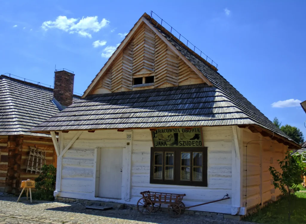Drewniana chata w skansenie w Sanoku z szyldem „Pracownia obuwia Jana Szudego”. Tradycyjna architektura drewniana z podwórkiem wyłożonym kamieniami.