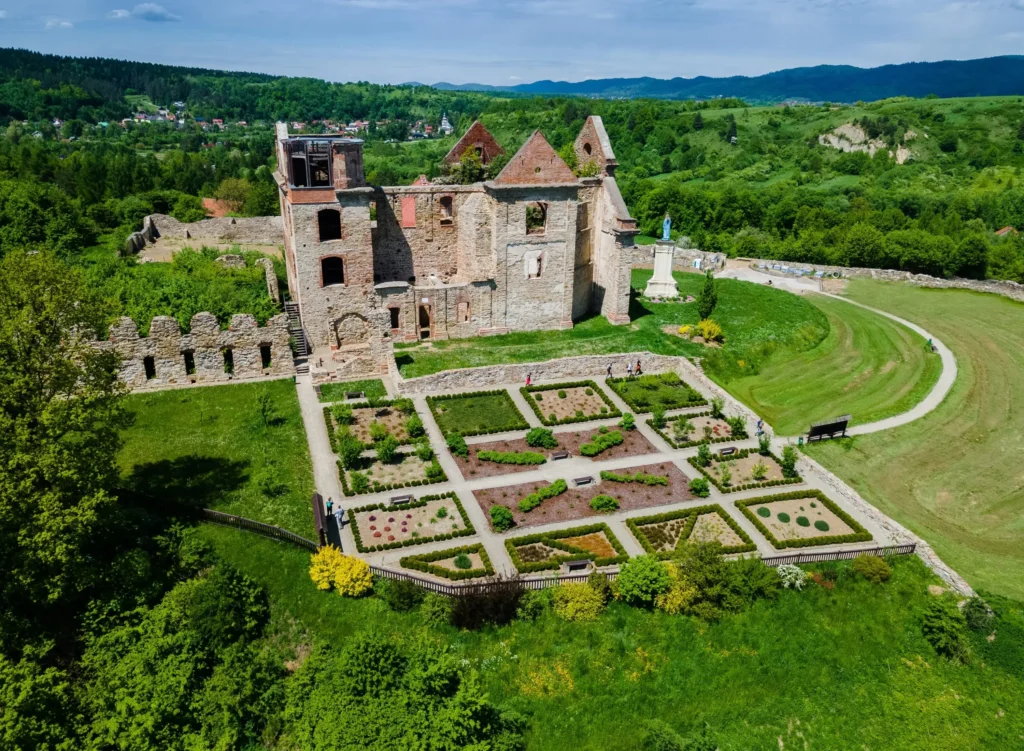 Ruiny Zamku Karmelitów w otoczeniu zieleni, z pięknie zaprojektowanym ogrodem na pierwszym planie. Malowniczy krajobraz gór i lasów w tle podkreśla historyczny urok tego miejsca.
