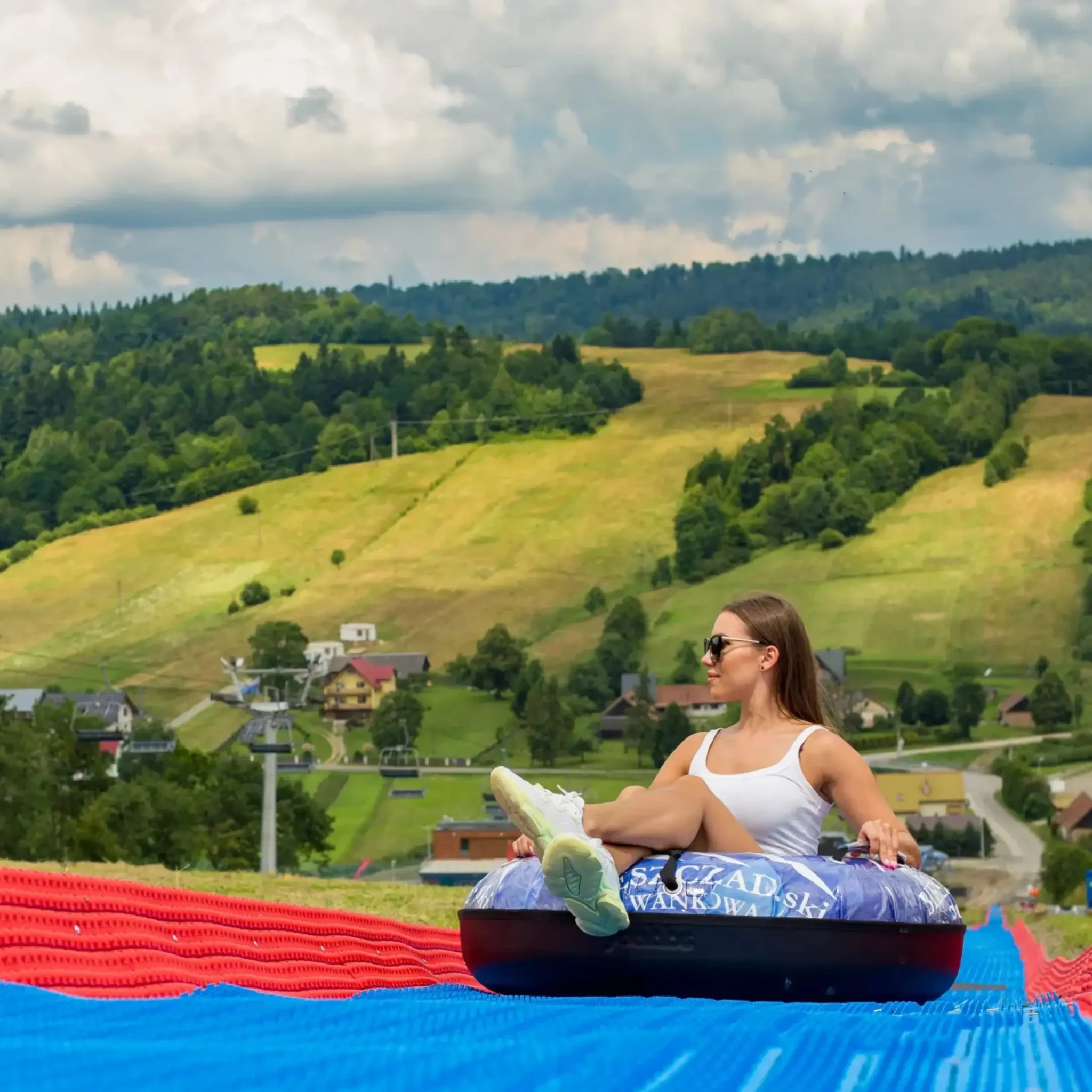 Kobieta zjeżdżająca na specjalnym pontonie po torze tubingowym z widokiem na zielone wzgórza Bieszczadów.