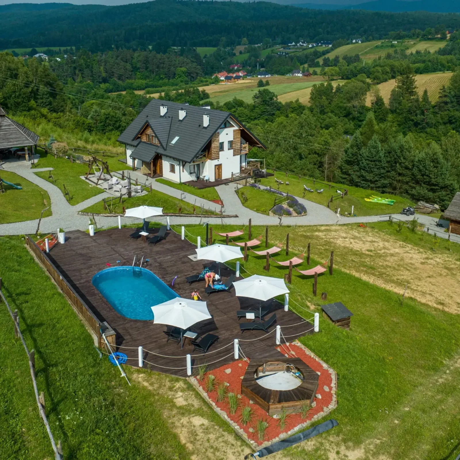 Widok z lotu ptaka na basen, leżaki i drewnianą chatę w Bieszczadach, z otaczającą zielenią i górami.