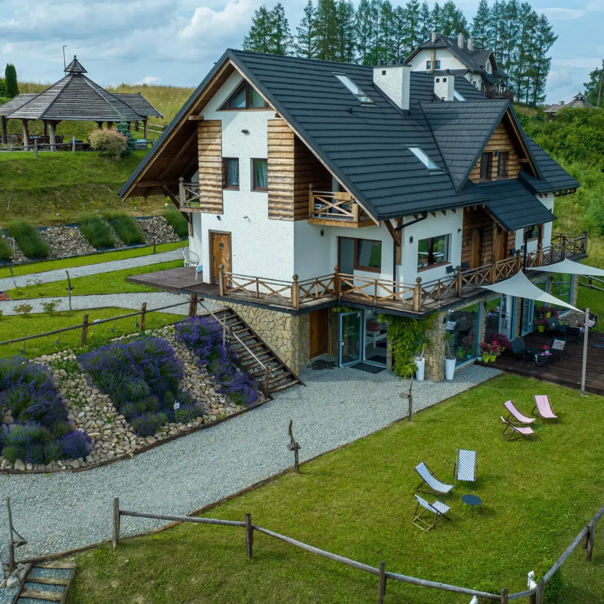 Widok na drewniany budynek z tarasem w Bieszczadach, otoczony ogrodem z lawendą i zielenią.