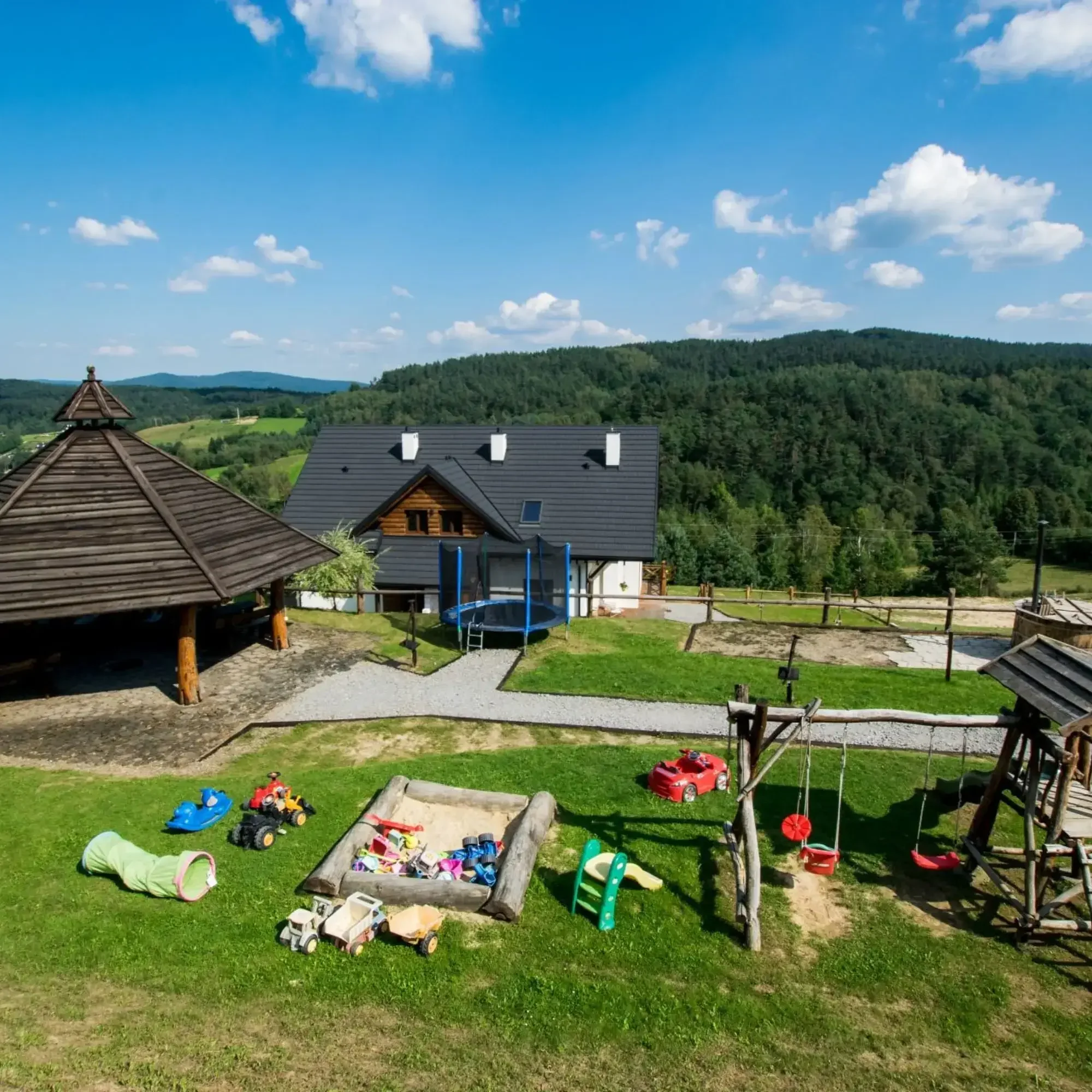 Obszar wypoczynkowy na świeżym powietrzu z placem zabaw dla dzieci, drewnianymi zabawkami i widokiem na góry i las w tle.