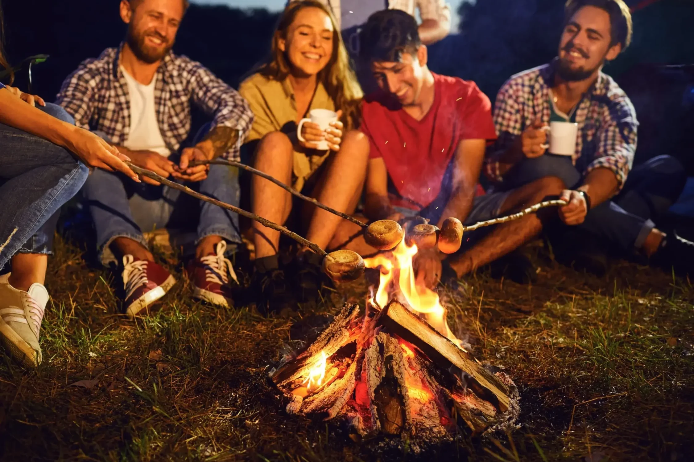 Grupa przyjaciół siedząca przy ognisku, piekąca jedzenie na patykach i ciesząca się wspólnym wieczorem.